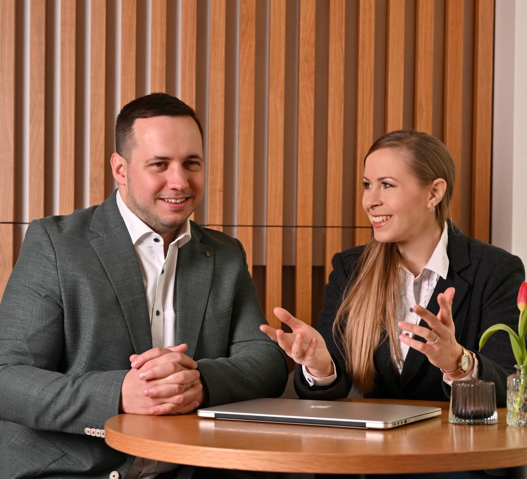 Mann und Frau im Business-Outfit sitzen an einem runden Holztisch, unterhalten sich freundlich und haben einen geschlossenen Laptop vor sich